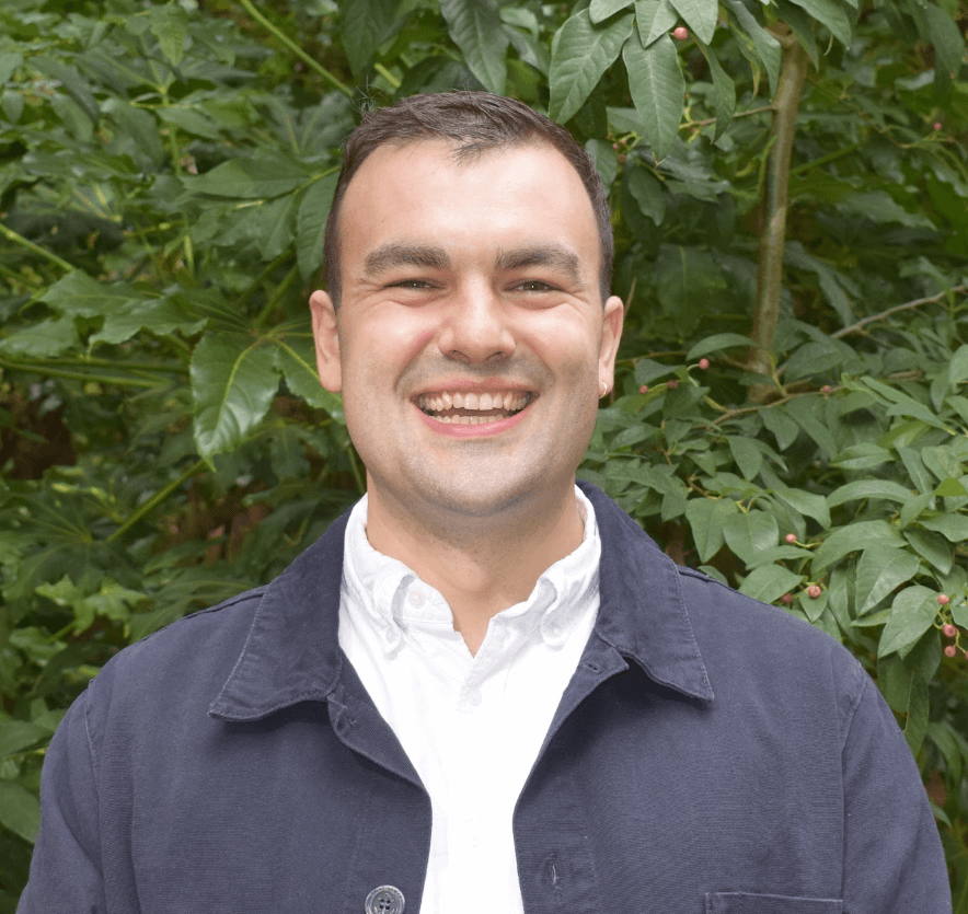 A picture of Money and Mental Health's Senior Research Officer Toby Murray, smiling while standing in front of a tree.