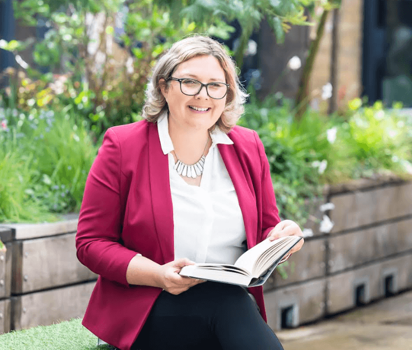 Photo of Jo Hill in a burgundy jacket smiling with an open book in her hand