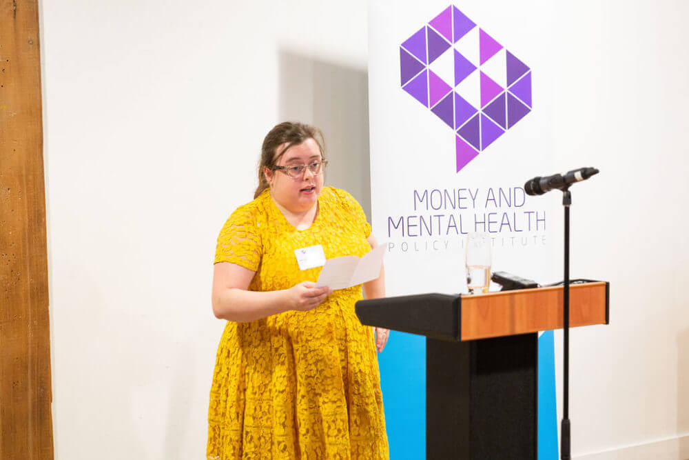 Helen Barker standing in front of podium and Money and Mental Health banner