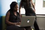 woman sitting down using a laptop with another woman standing over her
