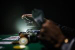 An image of a poker table, with some playing cards and poker chips on the table, and one person's hand holding poker chips with another holding playing cards.