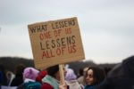 A woman holding a cardboard sign that reads, "What lessens one of us lessens all of us"
