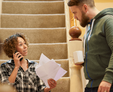 woman sitting on the stairs talking on the phone and holding bills in her hands with her partner standing up next to her