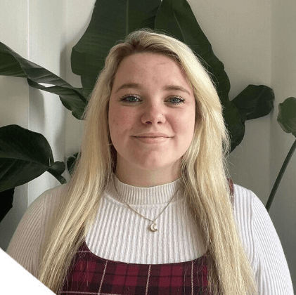 A photo of Cara MacSherry, standing indoors with a plant in the background.