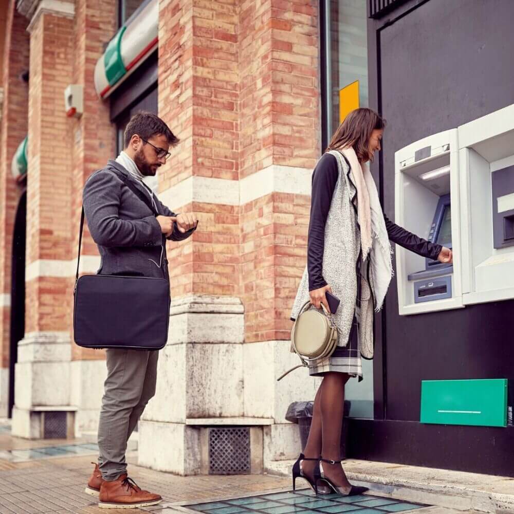 A person using an ATM with another person waiting behind them.