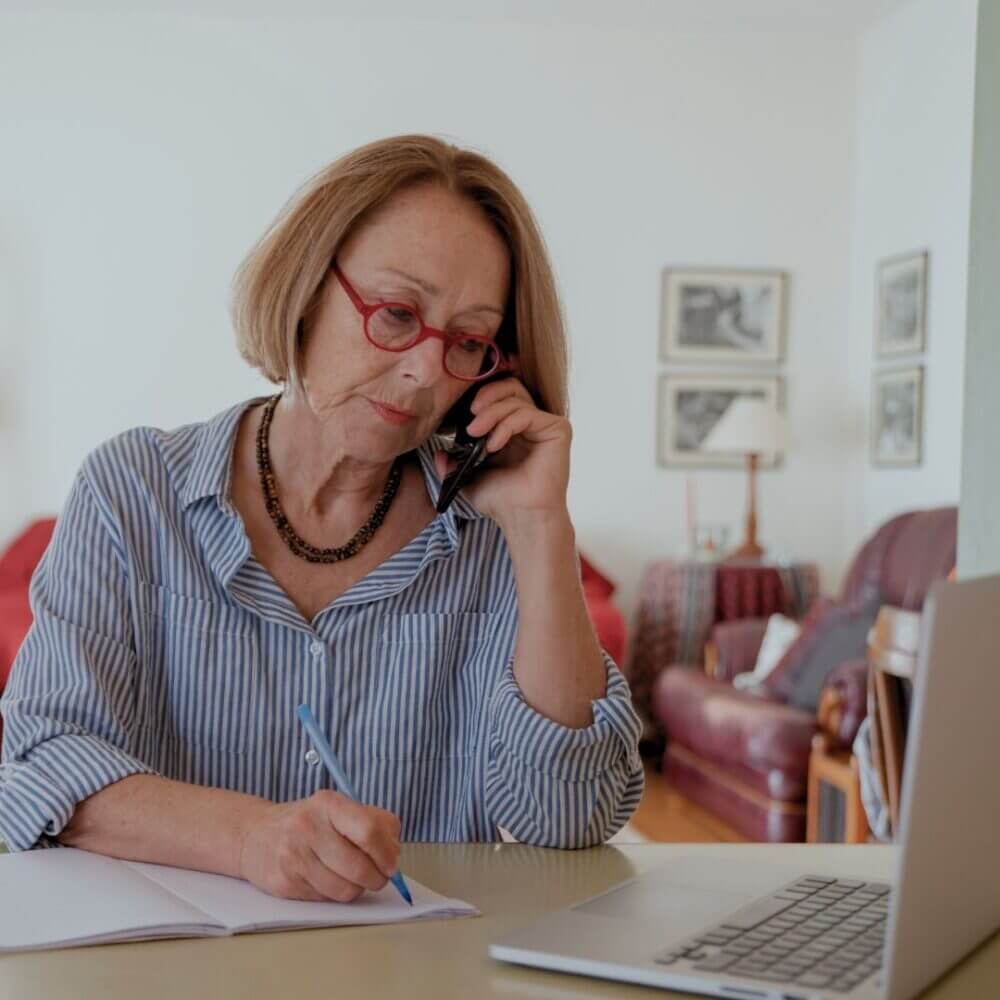 A person wearing red glasses, on the phone and sitting at a desk with a laptop, pen and paper.