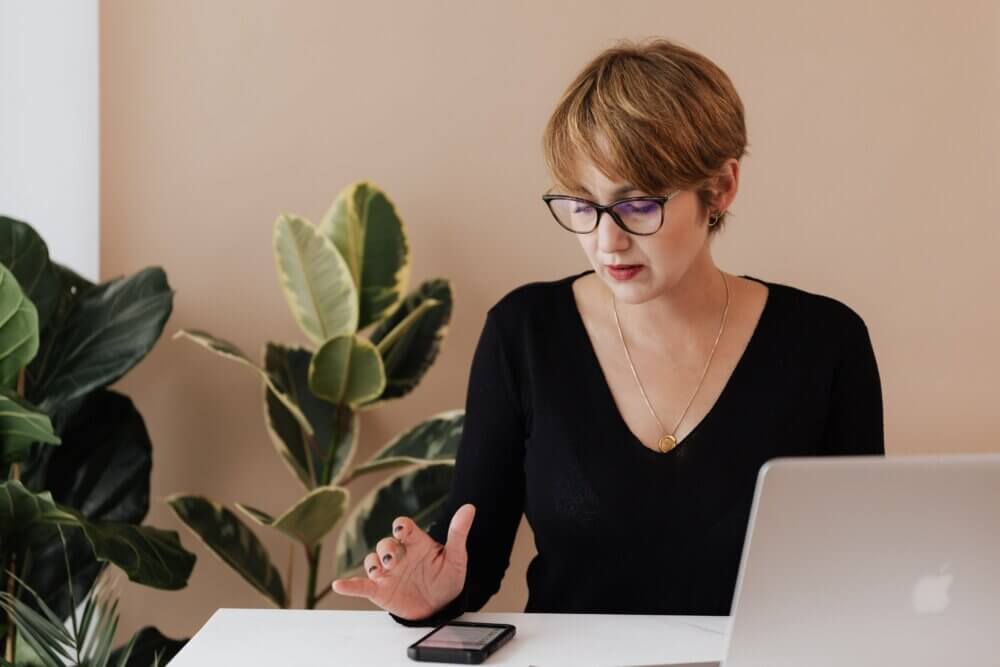 A photograph of a person looking at a mobile phone while sitting in front of a laptop.