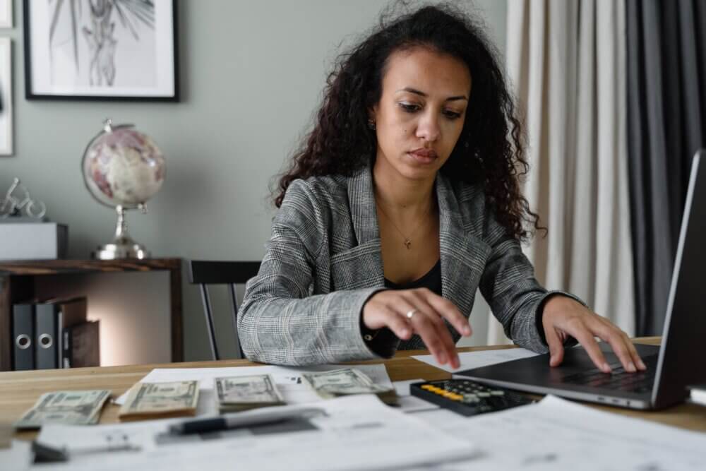 Image of woman using a laptop and calculator to perform calculations. She is sitting at a desk which is covered with paper and bank notes.