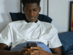 A man, sat in bed, looks glumly at his phone