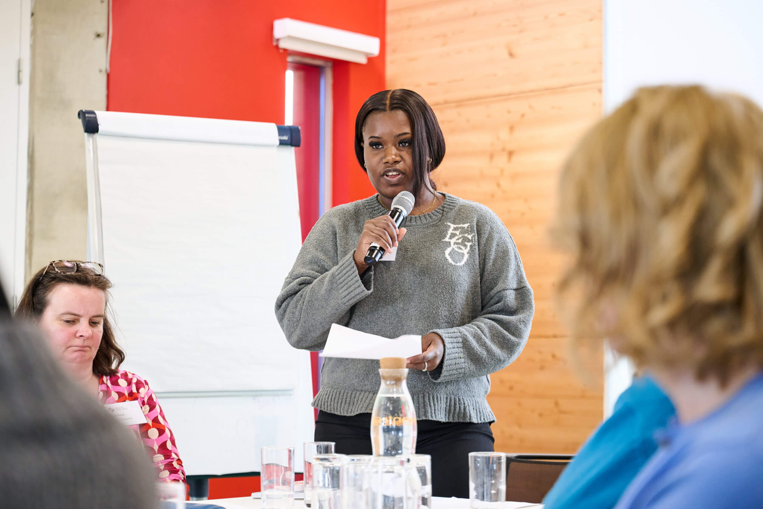People sat at a table, with one person presenting notes with a microphone - from our event focused on improving support services alongside members of our Research Community.