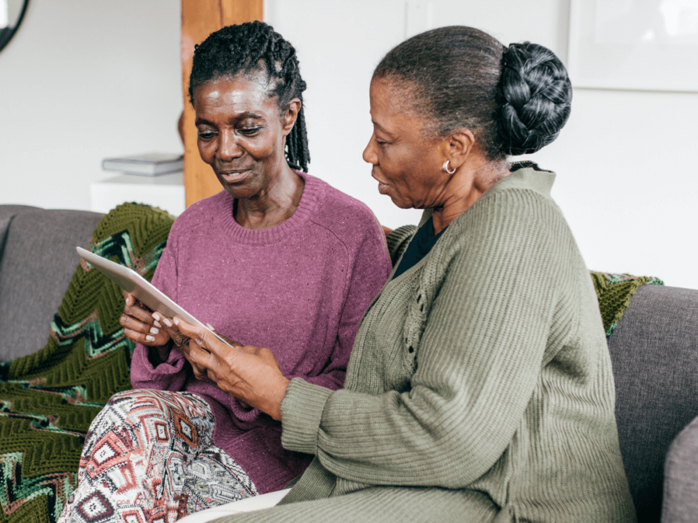 Two people, sat at home on a sofa looking at a tablet. One could be the other's carer supporting with finances.